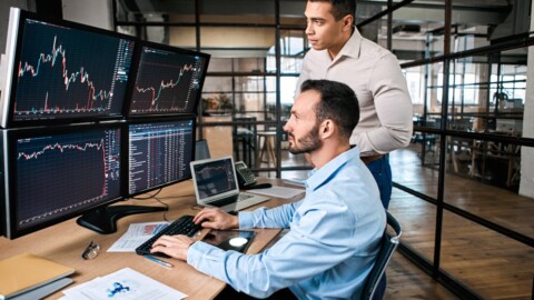 Two investors examine stock price data displayed on four screens as one investor sits and types into the computer.